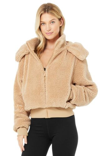 Photo of a blonde woman wearing black leggings and Alo Yoga's Foxy Sherpa zip-up jacket in camel